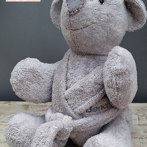 May include: A grey teddy bear made from soft fabric, with a heart-shaped patch on its nose. The bear is wearing a robe and has the word "Bench" embroidered on it. The bear is sitting on a wooden surface.