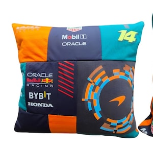 May include: A square decorative cushion featuring a patchwork design of Formula 1 racing team logos and colours. The fabric includes orange, dark blue, and teal panels with printed text such as Oracle, Red Bull Racing, Bybit, Honda, and the number 14.