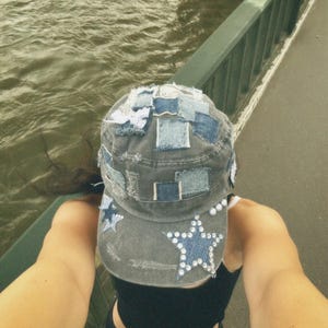 May include: A gray denim cap with a patchwork design and star embellishments. The cap is worn by a person with a black tank top and denim shorts.