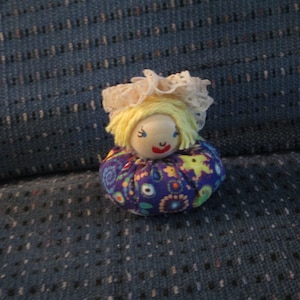 May include: A small, handmade doll with a yellow yarn wig, a white lace bonnet, and a colorful floral patterned dress.