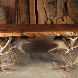 May include: A rustic wooden table with a live edge top, supported by intricately arranged deer antlers. The table's rich brown surface contrasts with the lighter tones of the antlers, creating a unique and natural aesthetic. A wooden barrel is in the background.
