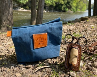 May include: A blue canvas pouch with a leather patch that reads "Fish Simply Fish Often" next to a leather fly holder with scissors. The items are on a bed of rocks and dirt, with a river and trees in the background.