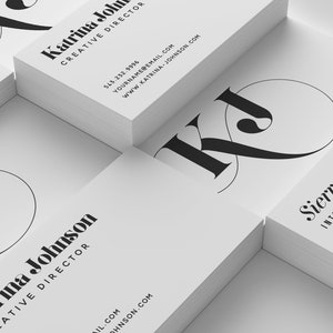 Puede incluir: Una pila de tarjetas de visita blancas con texto negro y un logotipo "KJ" estilizado. El texto de la tarjeta superior dice "Katrina Johnson, Directora Creativa". El texto de la tarjeta inferior dice "Sierra Rossi, Dise&ntilde;o de Interiores".