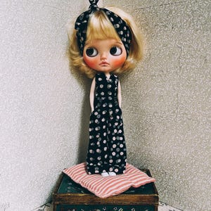 May include: A doll with blonde hair wearing a black and white polka dot jumpsuit and a black and white headband. The doll is standing on a pink and white striped cushion on top of a wooden box with a green painted lid.