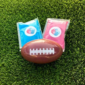 Peut inclure: Un ballon de football américain marron avec des rayures blanches est posé sur un gazon artificiel vert. Deux sachets en plastique scellés, un bleu et un rose, sont derrière le ballon. Chaque sachet a un autocollant blanc avec un motif rouge et bleu.