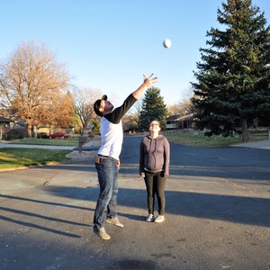 May include: A man and a woman are playing catch with a white ball in a residential neighbourhood. The man is wearing a white shirt with black stripes and blue jeans. The woman is wearing a grey sweater and black trousers.