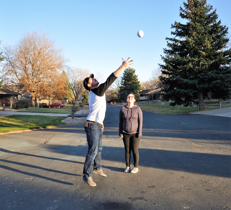 May include: A man and a woman are playing catch with a white ball in a residential neighbourhood. The man is wearing a white shirt with black stripes and blue jeans. The woman is wearing a grey sweater and black trousers.