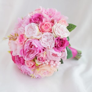 Puede incluir: Un ramo de flores redondo con un arreglo denso de rosas y peonías artificiales en varios tonos de rosa, desde el rubor pálido hasta el magenta intenso, acentuado con pequeñas hojas verdes y una cinta rosa envuelta en el mango.