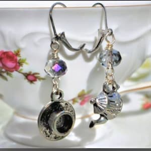May include: Silver-toned dangle earrings with a teacup and teapot charm. Each earring features a faceted crystal bead and a small charm. The earrings are displayed in front of a floral teacup.