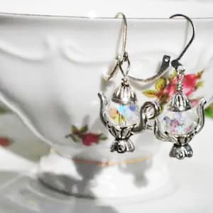May include: A pair of silver-toned teapot earrings with iridescent crystal-like centers. The earrings have a detailed design, including spouts, handles, and decorative bases. The background is a white teacup with floral patterns.