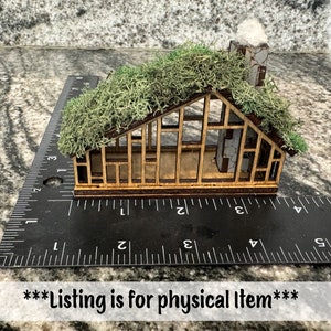 K&ouml;nnte beinhalten: Ein Miniatur-Holzhaus mit einem moosbedeckten Dach und einem Steinkamin. Das Haus ist etwa 7,6 cm lang und wird zur Ma&szlig;stabsbestimmung an einem Lineal platziert. Der Text "Goober Creations" befindet sich in der oberen rechten Ecke.