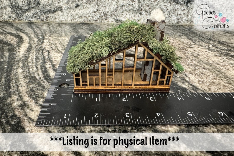 K&ouml;nnte beinhalten: Ein Miniatur-Holzhaus mit einem moosbedeckten Dach und einem Steinkamin. Das Haus ist etwa 7,6 cm lang und wird zur Ma&szlig;stabsbestimmung an einem Lineal platziert. Der Text "Goober Creations" befindet sich in der oberen rechten Ecke.