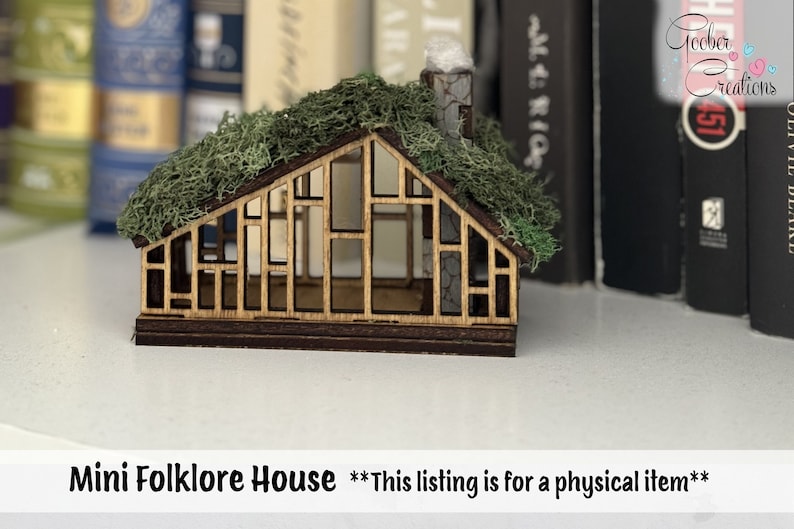 K&ouml;nnte beinhalten: Ein Miniatur-Holzhaus mit einem moosbedeckten Dach und einem Schornstein. Das Haus hat ein geometrisches Design mit mehreren Fenster&ouml;ffnungen. Der Text "Mini Folklore House" befindet sich am unteren Rand des Bildes.