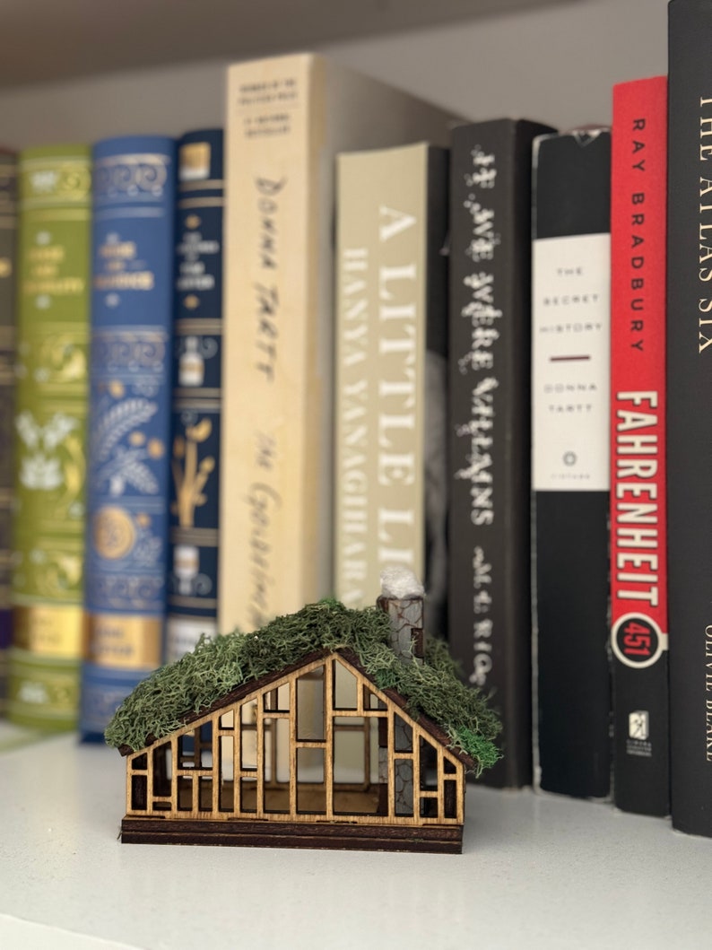 K&ouml;nnte beinhalten: Ein Miniatur-Holzhaus mit Moosdach und Schornstein, vor einer Reihe von B&uuml;chern platziert. Das Haus hat einen detaillierten Rahmen und ein Fensterdesign. Die B&uuml;cher tragen Titel wie "Fahrenheit 451" und "A Little Life".