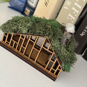 K&ouml;nnte beinhalten: Ein Miniatur-Gew&auml;chshaus aus Holz mit einem moosbedeckten Dach und einem kleinen Schornstein. Die Struktur hat einen dunkelbraunen Rahmen und einen hellbraunen Innenraum mit detailliertem Fensterdesign. Der Hintergrund umfasst eine Sammlung von B&uuml;chern.