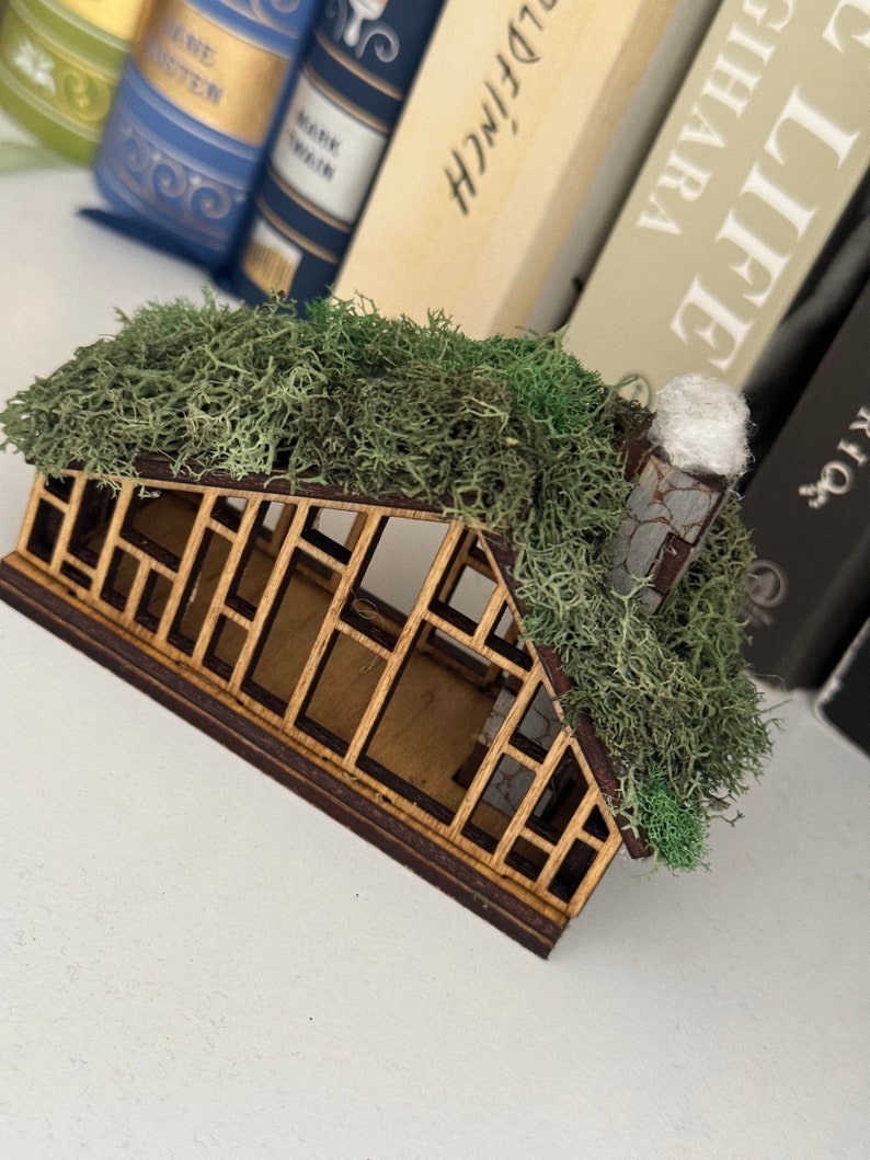 K&ouml;nnte beinhalten: Ein Miniatur-Gew&auml;chshaus aus Holz mit einem moosbedeckten Dach und einem kleinen Schornstein. Die Struktur hat einen dunkelbraunen Rahmen und einen hellbraunen Innenraum mit detailliertem Fensterdesign. Der Hintergrund umfasst eine Sammlung von B&uuml;chern.