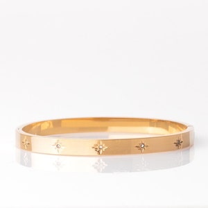 Może przedstawiać: Złota bransoletka typu bangle o polerowanym wykończeniu z powtarzającym się wzorem małych, gwiazdkowatych wzorów z przezroczystym kamieniem w środku.