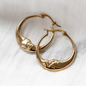 Op de afbeelding: Een paar gouden oorringen met een verhoogd, gebeeldhouwd ontwerp van een maansikkelgezicht op het onderste deel van elke ring. De oorbellen hebben een gepolijste, reflecterende afwerking en zijn voorzien van een scharnierend sluitmechanisme.