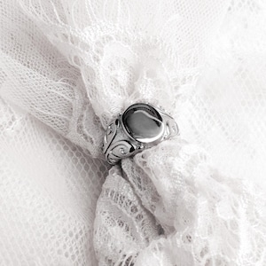 May include: A silver ring with an oval, unadorned center stone. The ring features ornate, swirling designs on the band. The ring is set against a backdrop of delicate, white lace.