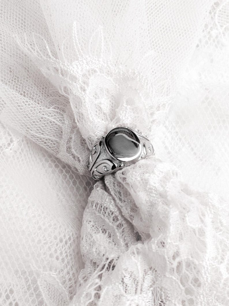 May include: A silver ring with an oval, unadorned center stone. The ring features ornate, swirling designs on the band. The ring is set against a backdrop of delicate, white lace.