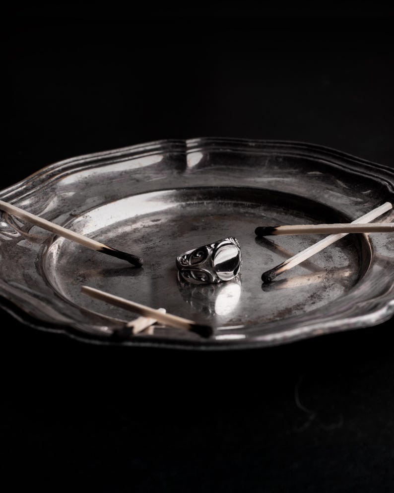 May include: A silver ring with an oval black stone rests on a tarnished silver plate, surrounded by burnt matchsticks. The ring features an ornate design. The plate has a scalloped edge, and the background is black.