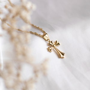 May include: A gold chain necklace with a gold cross pendant. The cross has a detailed design with a rounded centre.