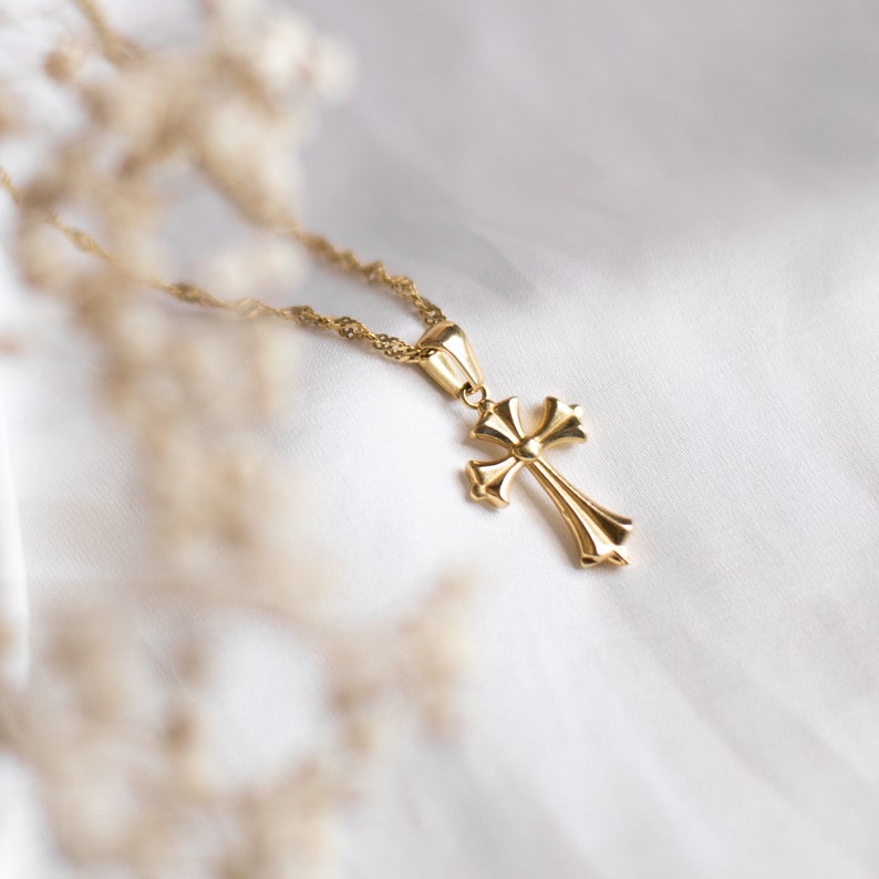 May include: A gold chain necklace with a gold cross pendant. The cross has a detailed design with a rounded centre.