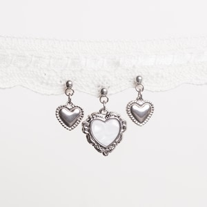 Op de afbeelding: Drie zilveren hartvormige hangers die aan een witte kanten rand hangen. Twee hangers hebben een eenvoudig hartontwerp, terwijl de middelste hanger een hartvormige parelmoer inleg heeft in een decoratief frame. De achtergrond is effen wit.
