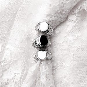 May include: Three silver rings with oval-shaped faces, one black and two white, are displayed on a white lace fabric. The rings have ornate, swirling designs around the face. The background is a delicate, textured white lace.