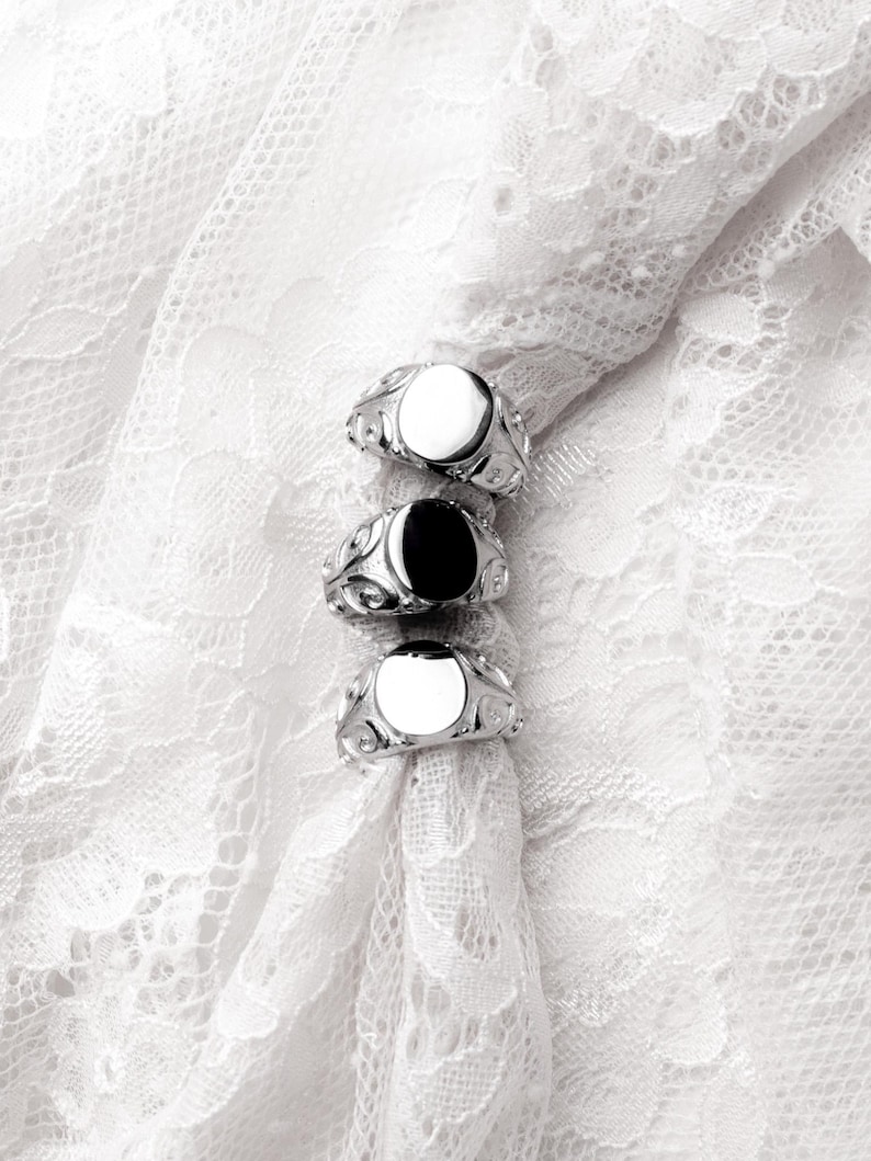 May include: Three silver rings with oval-shaped faces, one black and two white, are displayed on a white lace fabric. The rings have ornate, swirling designs around the face. The background is a delicate, textured white lace.