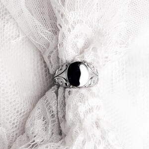 May include: A silver ring with an oval, black stone is set against a white lace background. The ring has an ornate design around the stone. The lace is delicate and adds a soft texture to the image. The ring is a piece of jewelry.