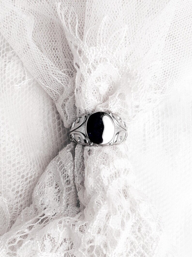 May include: A silver ring with an oval, black stone is set against a white lace background. The ring has an ornate design around the stone. The lace is delicate and adds a soft texture to the image. The ring is a piece of jewelry.