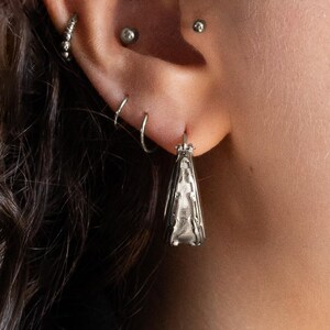 Op de afbeelding: Close-up van een zilveren oorbel met een gedetailleerd, langwerpig ontwerp. De oorbel hangt aan een doorboord oor, met meerdere piercings met kleine zilveren hoepels en studs. De vorm van de oorbel is een gestileerde, langwerpige driehoek.