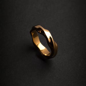 Op de afbeelding: Een goudkleurige ring met een golvend, getextureerd ontwerp. De ring heeft een gepolijst, reflecterend oppervlak en staat tegen een donkere achtergrond. De unieke vorm en kleur van de ring maken het een opvallend sieraad.