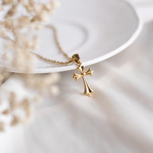 May include: A gold cross pendant necklace on a white surface. The cross has a detailed design with a rounded centre.