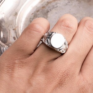 May include: A silver signet ring with a blank, oval face. The ring features decorative detailing around the face and on the band. The ring is shown on a hand, with a silver plate in the background.