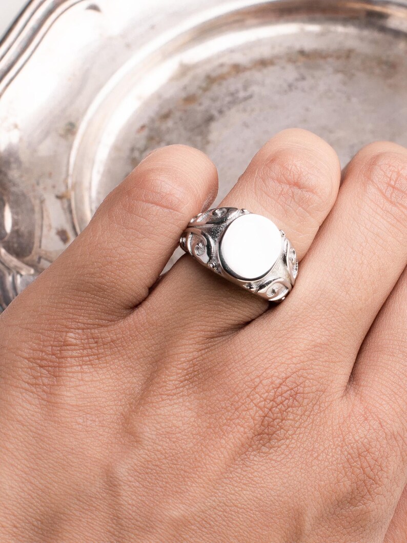May include: A silver signet ring with a blank, oval face. The ring features decorative detailing around the face and on the band. The ring is shown on a hand, with a silver plate in the background.