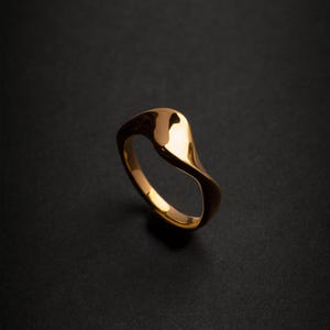 Op de afbeelding: Een goudkleurige ring met een glad, gebogen ontwerp. De ring heeft een gepolijste afwerking, die licht reflecteert. De band buigt rond om een doorlopende lus te vormen. De ring staat tegen een donkere achtergrond.