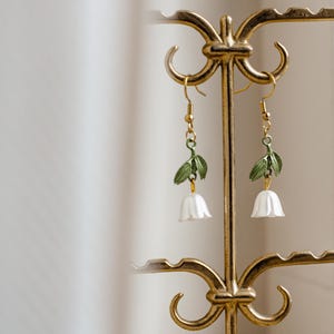 Op de afbeelding: Goudkleurige oorbellen met een bloemmotief. Elke oorbel heeft een witte, klokvormige bloem, groene bladeren en een gouden haak. De oorbellen worden tentoongesteld op een decoratieve gouden standaard.