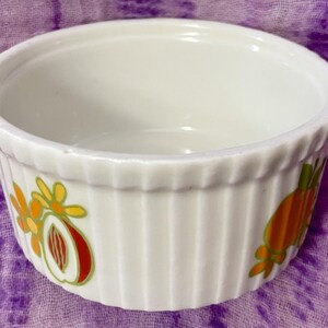 May include: White ceramic ramekin with vertical ribbed sides and a scalloped rim. The ramekin is decorated with colorful fruit and floral designs in orange, yellow, and red. A charming addition to any kitchen.