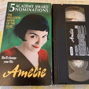 May include: A VHS tape of the movie Amélie, featuring a green cardboard sleeve with a portrait of a person with dark hair and red lipstick, the text 5 Academy Award Nominations, The feel-good movie of the year, She'll change your life, and Amélie.
