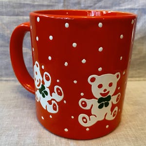 May include: A red ceramic mug with white polka dots and two white teddy bear illustrations. The bears have green bow ties. The mug has a curved handle and is suitable for hot or cold beverages.