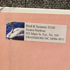 May include: A pink envelope with a white label featuring a swan image and text. The label includes the names "Brad & Suzanne TESH", "Swann Harbour", and an address in Swansboro, NC.
