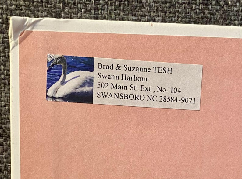 May include: A pink envelope with a white label featuring a swan image and text. The label includes the names "Brad & Suzanne TESH", "Swann Harbour", and an address in Swansboro, NC.