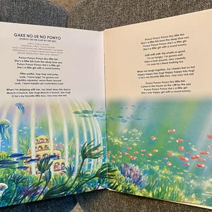 May include: An open book with colorful illustrations and text from "Ponyo on the Cliff by the Sea." The artwork depicts an underwater scene with fish, jellyfish, and a building. The text is in English and Japanese.