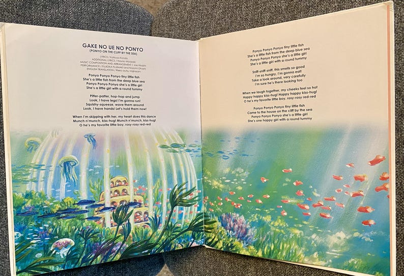 May include: An open book with colorful illustrations and text from "Ponyo on the Cliff by the Sea." The artwork depicts an underwater scene with fish, jellyfish, and a building. The text is in English and Japanese.