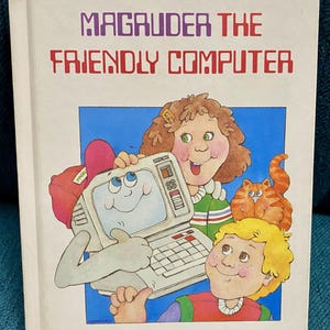 May include: A hardcover book titled "Magruder the Friendly Computer." The cover illustration features a cartoon computer with a smiling face, held by two children. A cartoon cat is also present. The title is in purple and red block letters.
