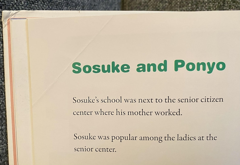 May include: A page from a book with the title "Sosuke and Ponyo" in green text. The text below describes Sosuke's school and his popularity at the senior center. The page has a cream-colored background.