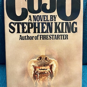 May include: A paperback book titled "CUJO" by Stephen King. The cover features a menacing dog's face with exposed teeth and drool. The title is in large black letters, with the author's name and "Author of FIRESTARTER" below.