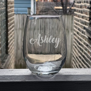May include: A clear glass stemless wine glass with the name "Ashley" etched on the side.